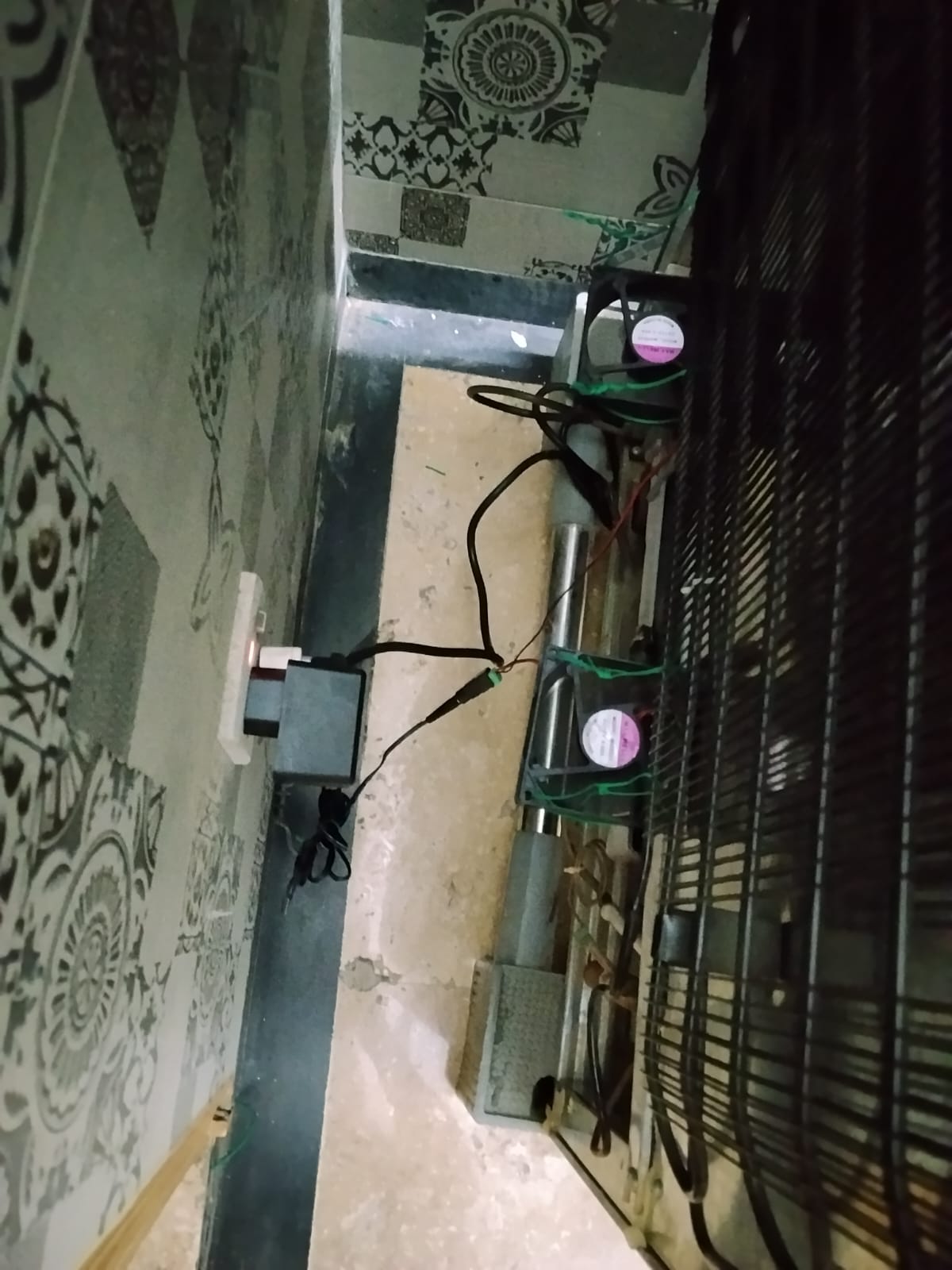Two small 12V DC cooling fans installed behind a domestic refrigerator, wired through a 12V power adapter to a wall socket.